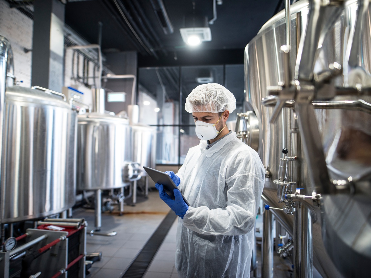 A man wearing a hair net, a mask, latex gloves, and a white jacket holds a tablet in front of large industrial tanks.
